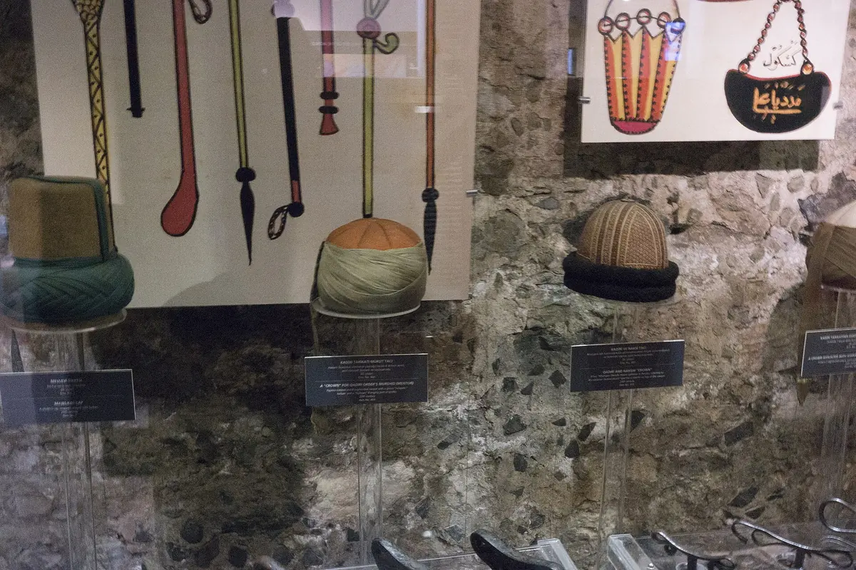 Filcowe kapelusze w kilku kolorach wyeksponowane na kamiennej ścianie obok zadrukowanych paneli w muzeum.